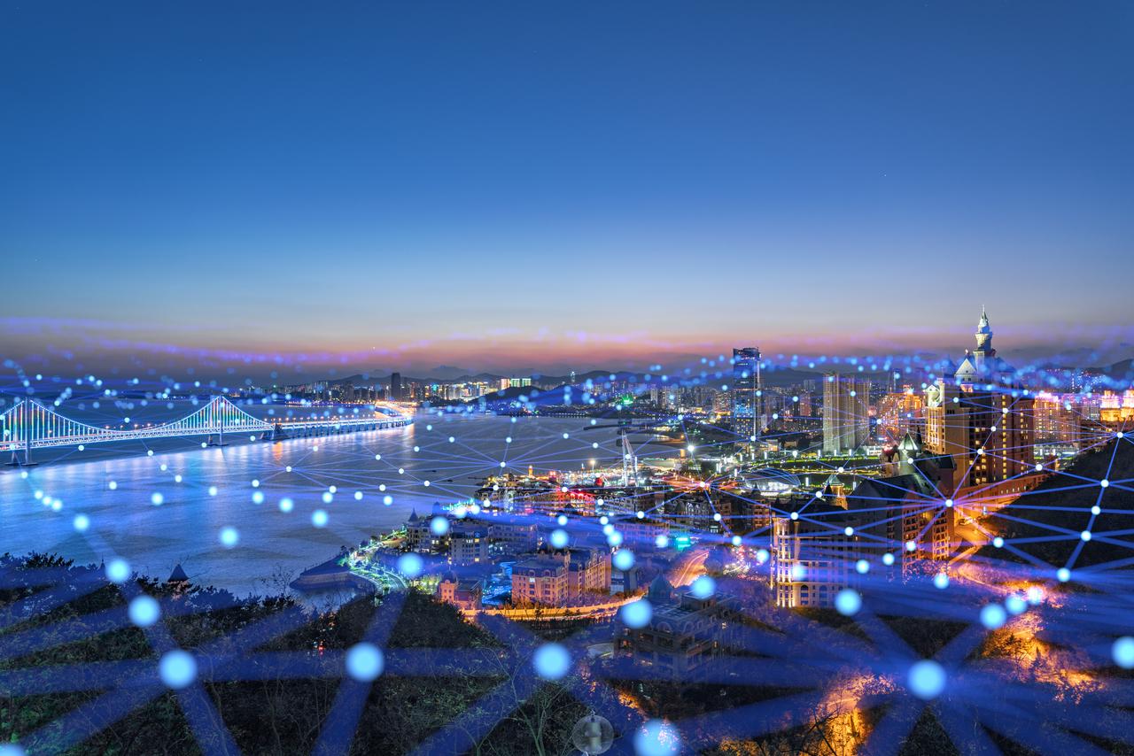 Panorámica nocturna de una ciudad costera moderna, destacando un puente iluminado que conecta ambos lados de un río. En el cielo y el agua, una red de puntos y líneas digitales simboliza la conectividad y la tecnología de la Industria 4.0. Edificios altos y modernos resaltan en el horizonte, mostrando un entorno urbano avanzado e interconectado. Imagen para la entrada del blog 'Espacios de Datos: La nueva clave de la Competitividad Industrial' por Telefónica España