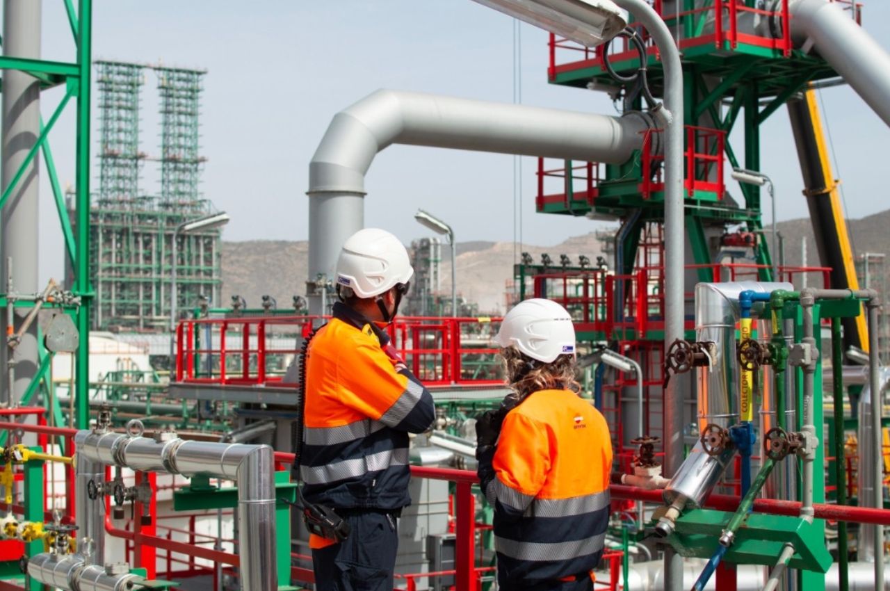 Dos operarios con cascos y chalecos reflectantes inspeccionando las instalaciones de una refinería de Repsol transformada en centro multienergético para la producción de combustibles renovables, un pilar de la autonomía estratégica industrial en España.